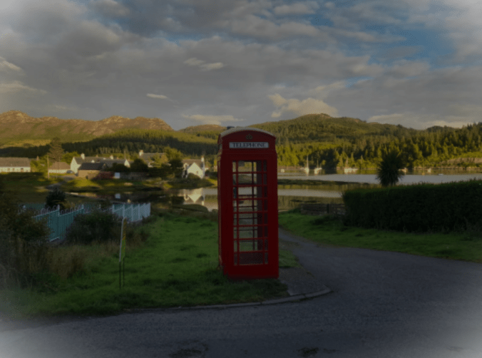 Red Phone booth overlooking a lush country village with a marina and a few boats, mountains and blue sky in the background.