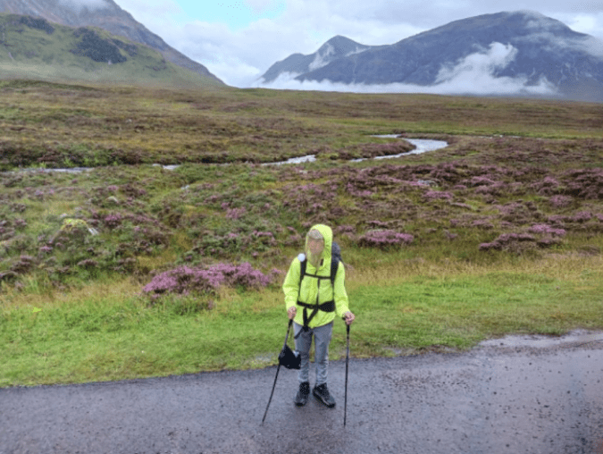 Woman with hiking poles, standing at the side of a road, tall mountains across a field and creek behind her.