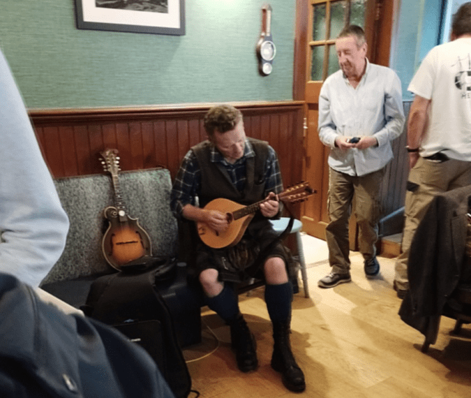Youndg man in kilt and knee socks on the bench playing a small guitar, spare by his side, in the hallway of a brightly lit modern pub.