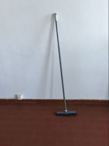 tall broom leaning against a flat white wall, flooring is polished brick.
